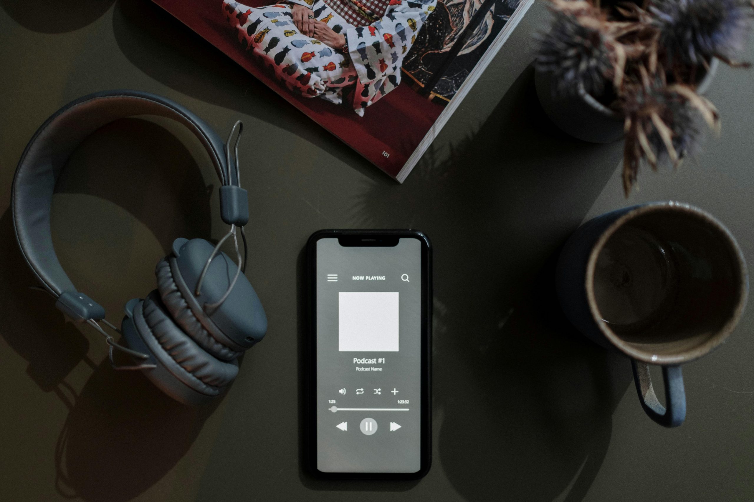A flat lay image of a smartphone playing a podcast next to headphones and a mug.